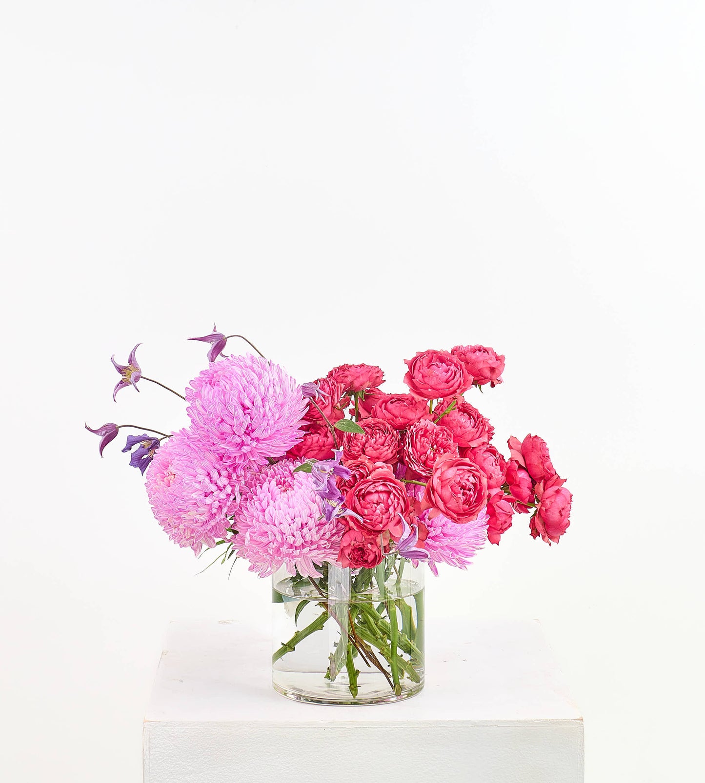 A vibrant floral arrangement featuring pink chrysanthemums, pink roses, and small purple star-shaped flowers in a clear glass jar vase, displayed on a white pedestal against a white background.