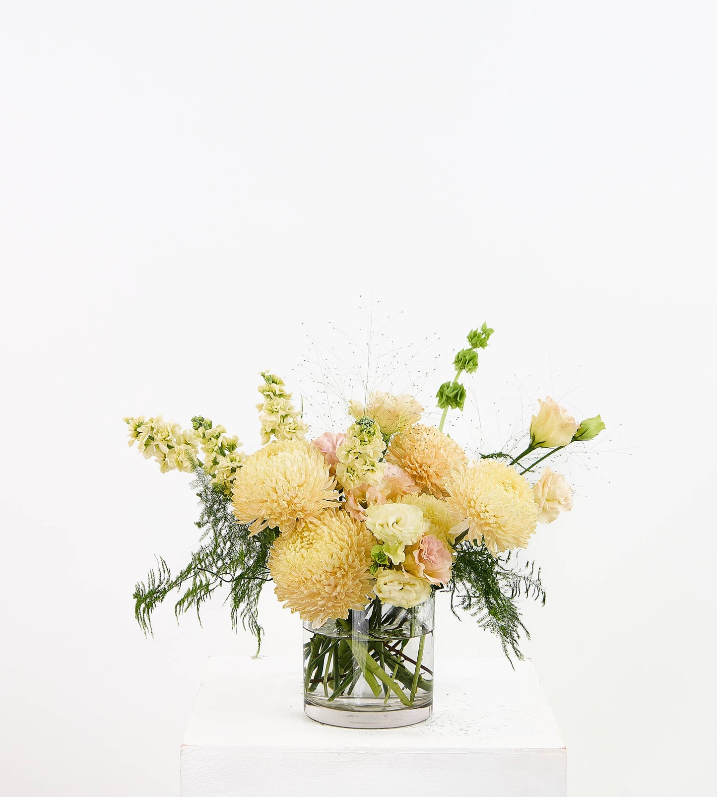 A floral arrangement of pale yellow, peach, and cream flowers with green foliage in a clear glass vase, displayed on a white pedestal against a white background.