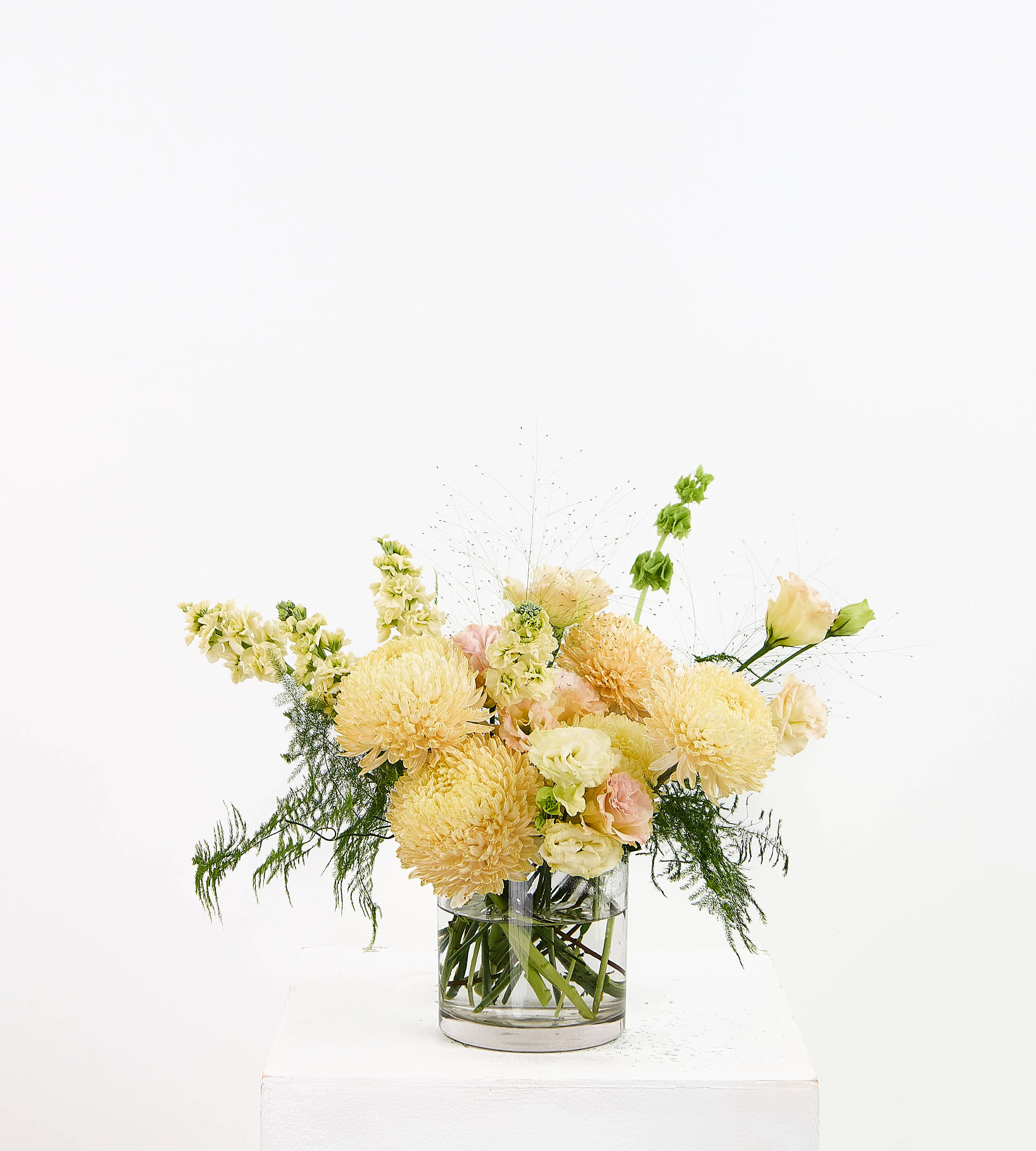 A floral arrangement of pale yellow, peach, and cream flowers with green foliage in a clear glass vase, displayed on a white pedestal against a white background.