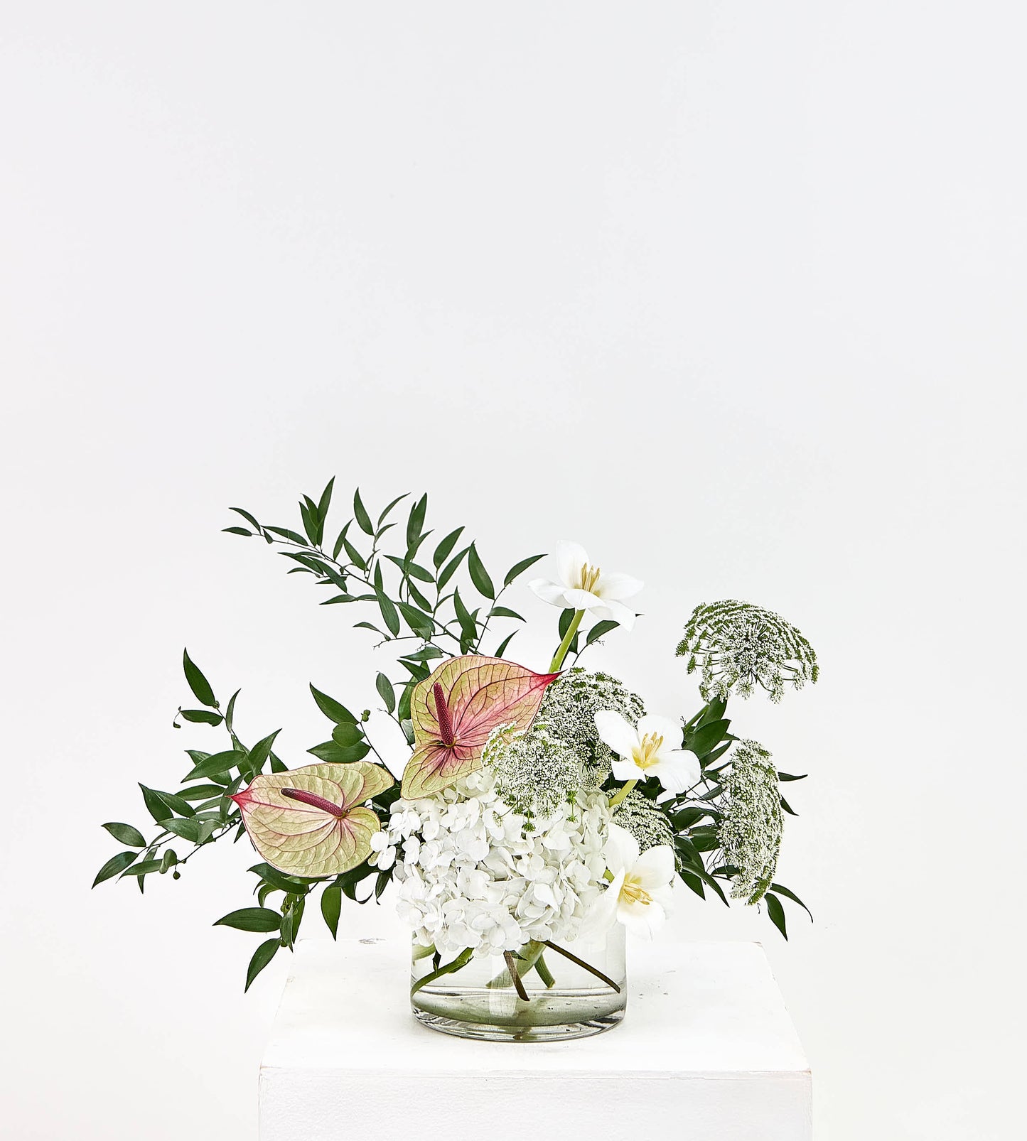 A floral arrangement featuring white flowers, green foliage, and pinkish-red anthuriums in a clear glass vase, displayed on a white pedestal against a white background.