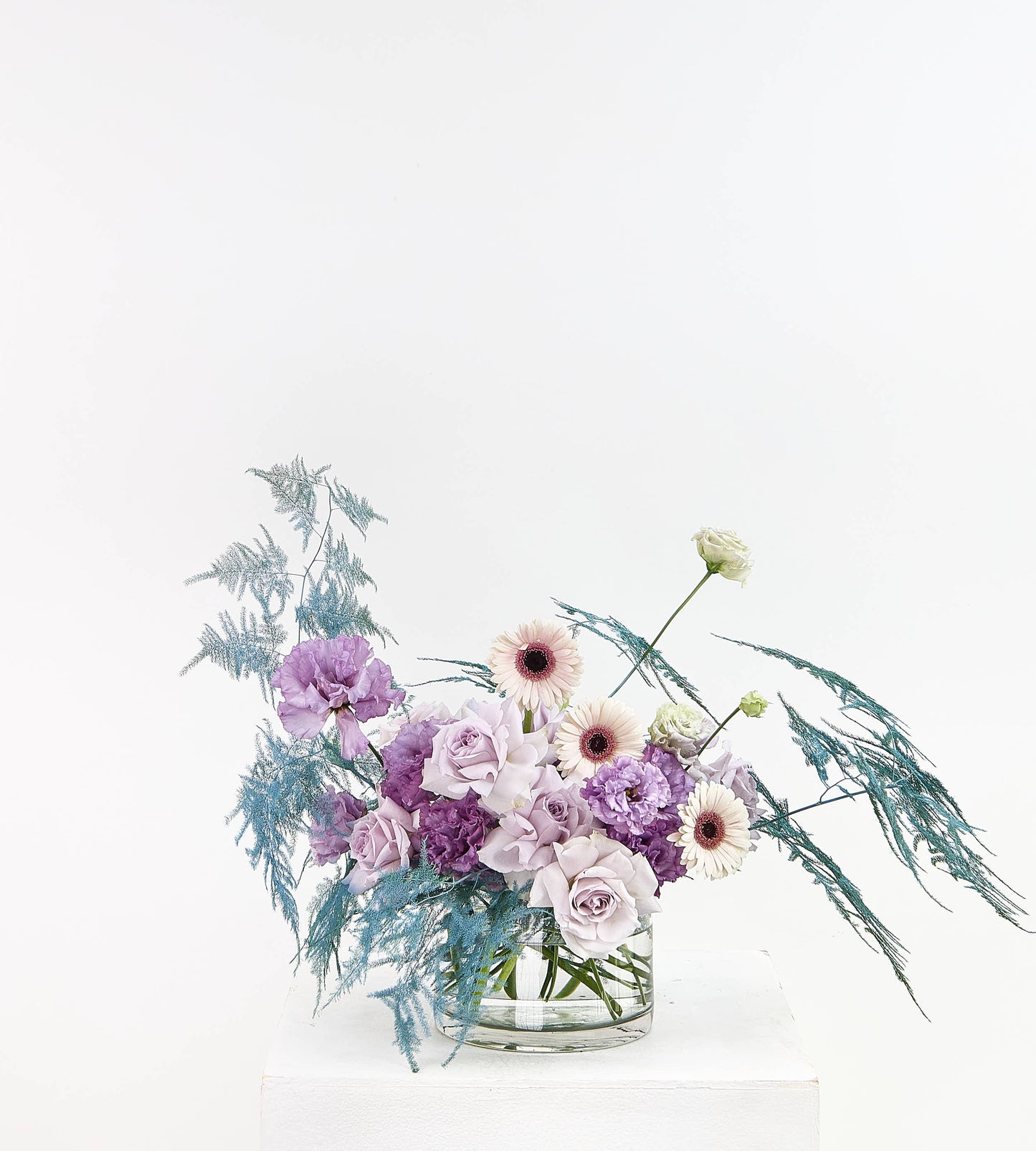 A floral arrangement in a clear glass vase on a white pedestal against a white background. The bouquet features light purple and lavender roses, white daisies with dark centers, and blue-green feathery filler stems.