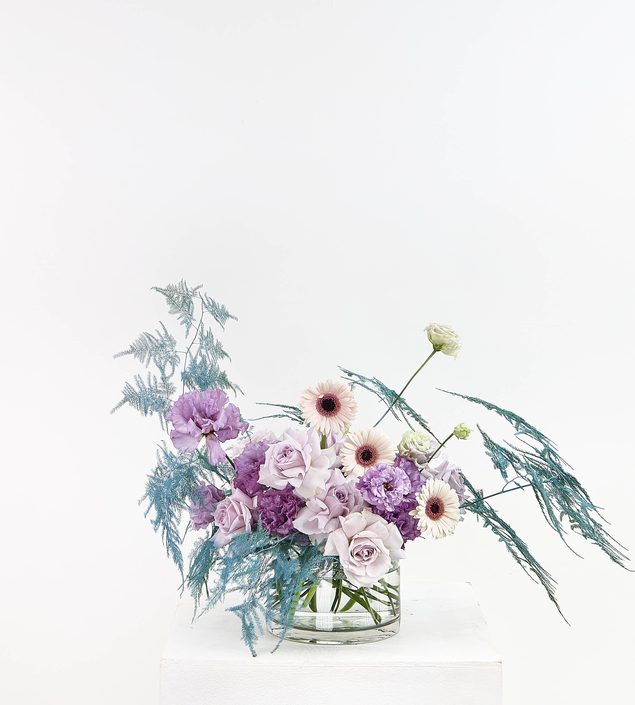A floral arrangement in a clear glass vase on a white pedestal against a white background. The bouquet features light purple and lavender roses, white daisies with dark centers, and blue-green feathery filler stems.